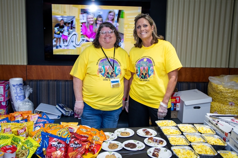 Two volunteers wearing matching yellow “Camp Read-A-Lot” literacy event shirts stand behind a snack table filled with popcorn, chips, cookies, pastries, and drinks. A display screen behind them shows photos of students reading and participating in literacy activities, highlighting a school event that celebrates books and reading.
