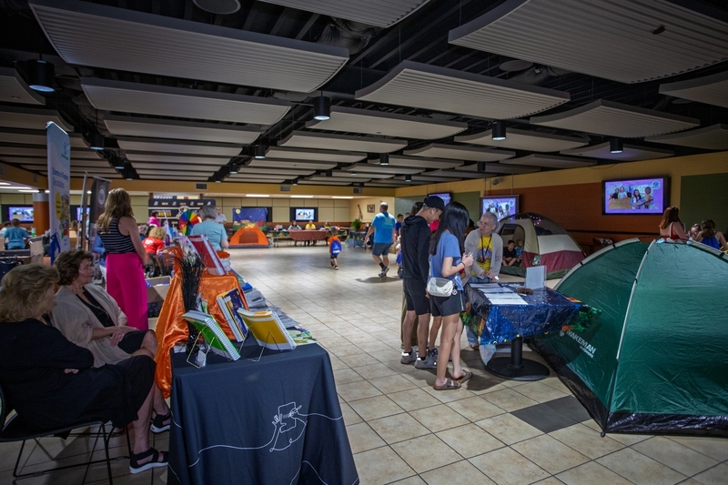 Large indoor literacy event with families, students, and community organizations gathered around activity tables, book displays, and camping themed reading stations. Several tents are set up throughout the room while attendees explore exhibits and resources, creating a festival atmosphere focused on reading, learning, and family engagement.