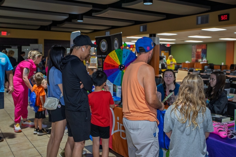Families visit literacy and community resource booths during a school reading event while volunteers share information and activities with children. A colorful prize wheel and tables filled with educational materials create an interactive atmosphere that encourages family engagement and participation in literacy programs.