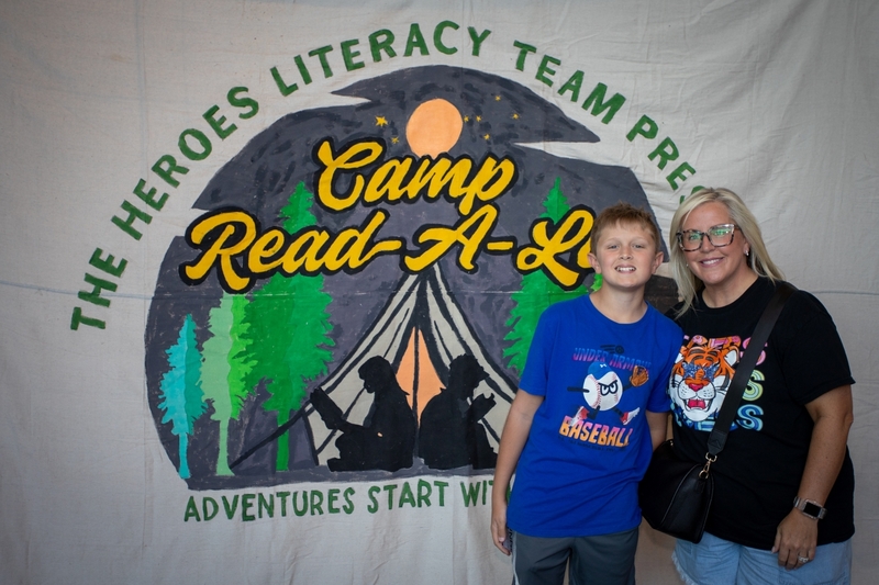 The Heroes Literacy Team Presents Camp Read A Lot" backdrop featuring a camping tent, trees, and readers gathered around a campfire. The display promotes a family literacy event with the theme "Adventures Start With Reading" and celebrates reading as a shared adventure.