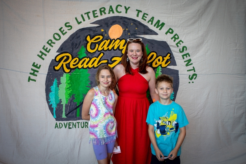 Woman in a red dress poses with two children in front of a "The Heroes Literacy Team Presents Camp Read A Lot" backdrop featuring a campsite, trees, and moonlit reading scene. The family smiles for a photo during a literacy event celebrating reading adventures and family engagement.