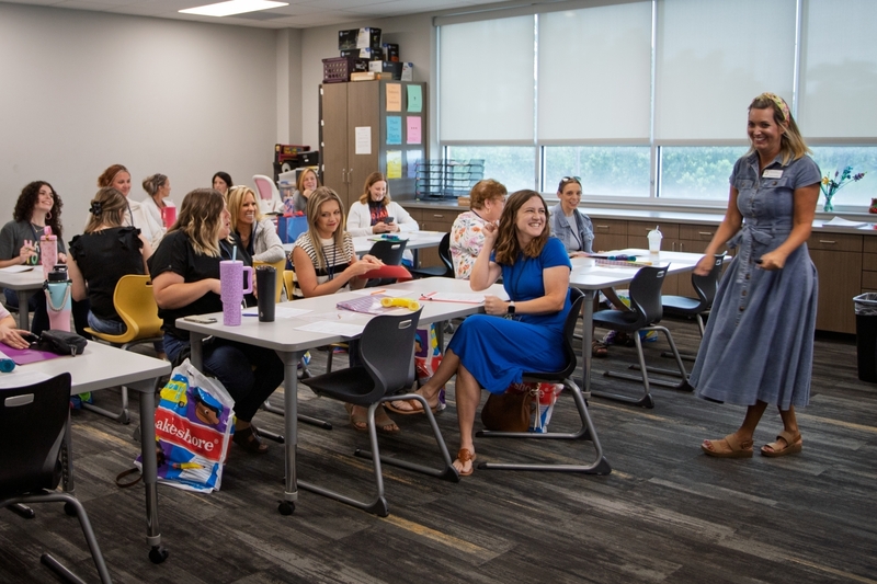 Educators sit at tables during a professional learning session while a presenter in a blue dress leads an interactive activity at the front of the classroom. Participants smile, engage in discussion, and follow along with materials spread across the tables in a bright training space.