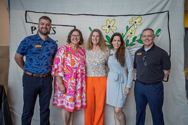 Five school leaders and educators stand together smiling in front of a decorative Pages Across the Plains backdrop featuring an open book and flowers. The group poses for a commemorative photo at an education and literacy event, wearing name badges and professional attire.