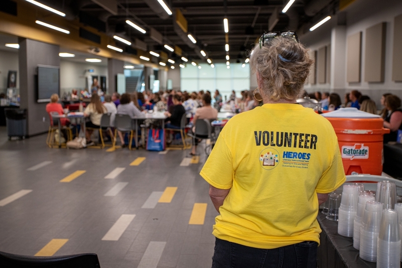 A volunteer wearing a yellow Heroes Literacy Instruction Team shirt stands near a refreshment station while a large group of educators attends a training session in the background. The back of the shirt reads "VOLUNTEER" and highlights the support role volunteers play during professional learning events.