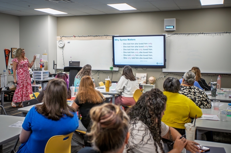 Educators sit in a professional development workshop while a presenter leads a lesson on sentence structure and meaning. A presentation slide titled "Why Syntax Matters" displays examples showing how emphasis changes when the word "only" is moved within a sentence, while attendees take notes and follow along.