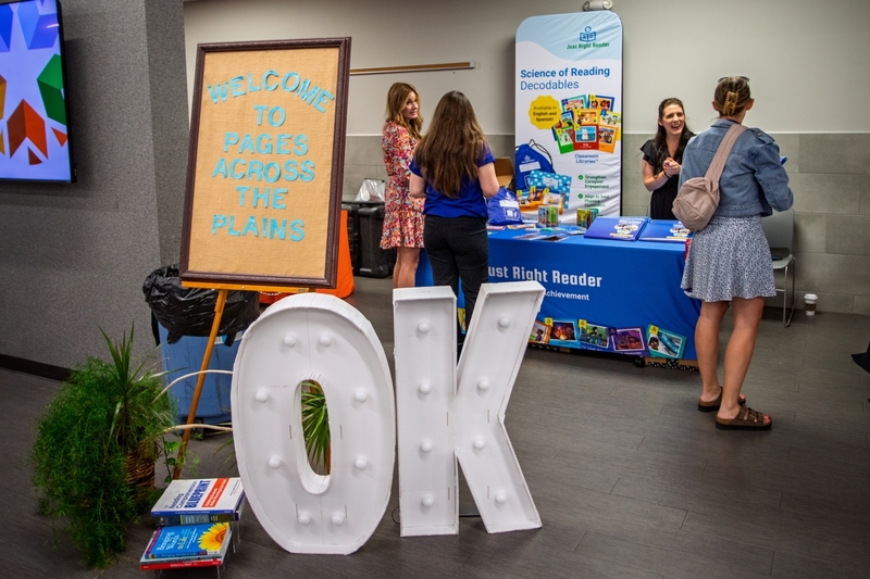 Display at a literacy conference featuring a sign that reads "WELCOME TO PAGES ACROSS THE PLAINS" beside large illuminated letters spelling "OK." Educators visit a Just Right Reader booth in the background showcasing Science of Reading decodable books and literacy resources for schools and classrooms.