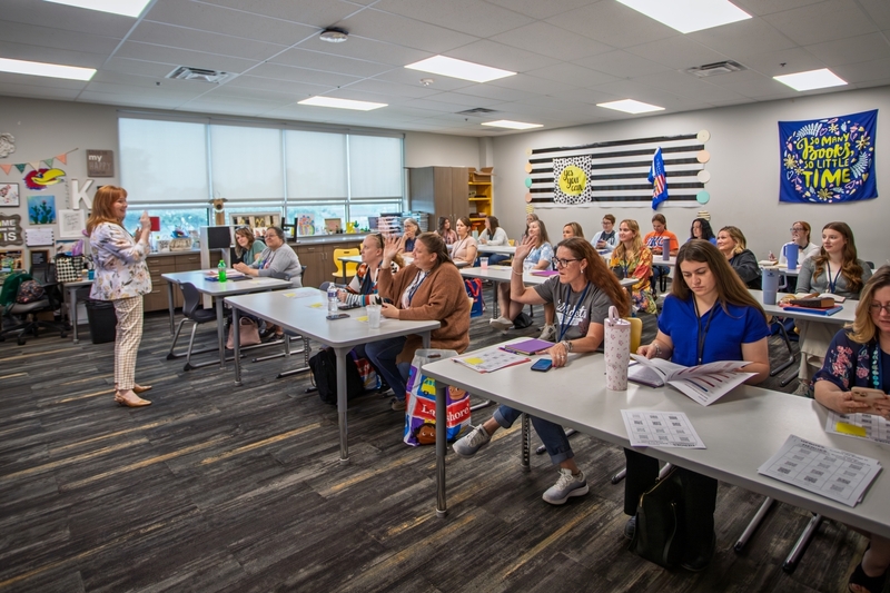 A presenter stands at the front of a classroom leading a professional development session while educators seated at tables raise their hands to participate. Training materials, notebooks, and drinks are spread across the tables as attendees engage in discussion and learning in a brightly decorated classroom.