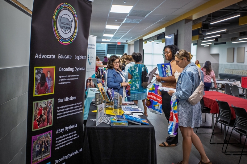 Attendees browse educational resources and speak with exhibitors at a Decoding Dyslexia information booth during a literacy conference. A large banner reads "Advocate. Educate. Legislate." while visitors review books, brochures, and materials focused on dyslexia awareness and literacy support.