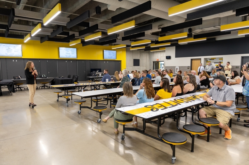 A speaker presents to a large audience seated at long cafeteria tables in a modern room with yellow wall accents and overhead acoustic ceiling panels.