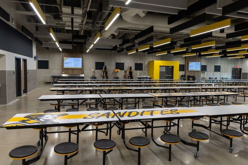 An empty modern school cafeteria filled with long grey and black tables, yellow-accented seating, and acoustic ceiling baffles, with the word "Sandites" printed on the tabletops.