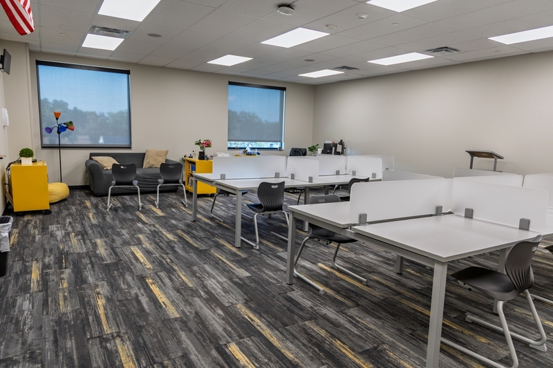A modern, open-plan classroom equipped with modular student desks, privacy dividers, grey chairs, and pattern-carpeted floors, with a sofa and yellow storage cabinets in the background.