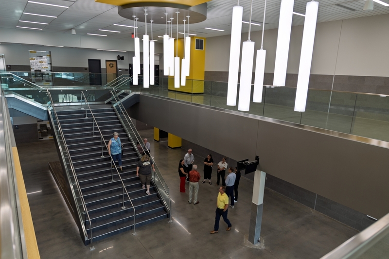 A high-angle view of a modern two-story school atrium featuring a wide central staircase, hanging cylindrical light fixtures, and a group of people conversing on the ground floor.