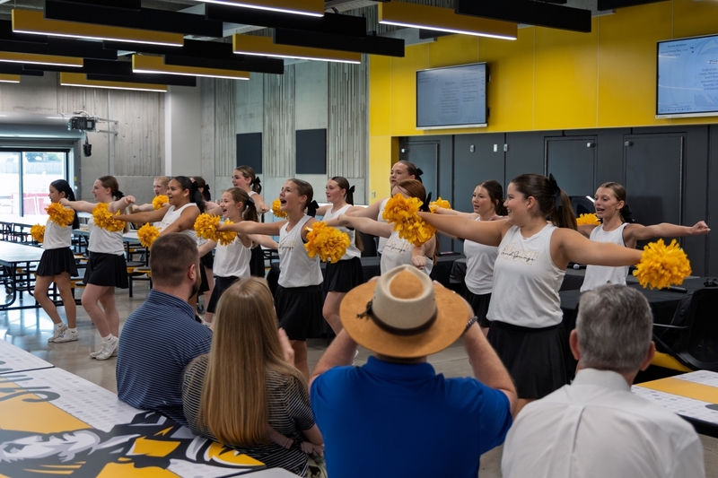 A group of cheerleaders in white sleeveless tops and black skirts perform a routine with gold pom-poms extended while an audience watches from foreground tables.