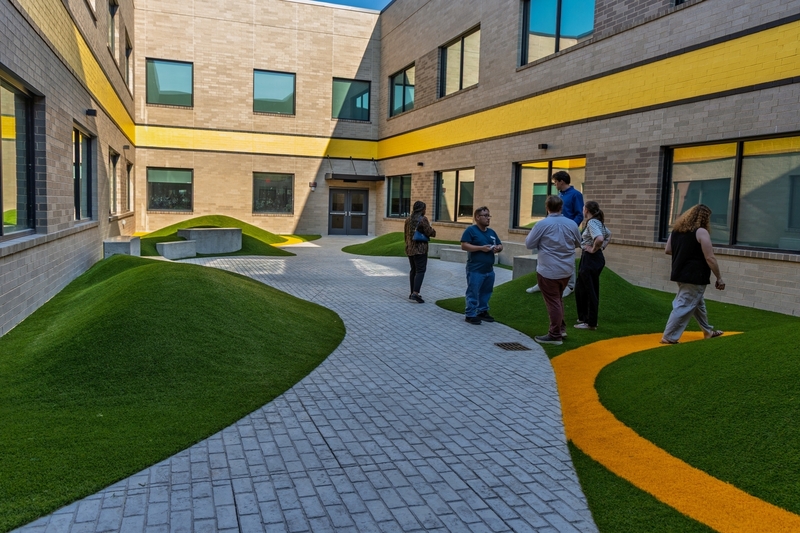 An outdoor courtyard with a gray paved walkway, small rolling mounds of green synthetic turf, and yellow accent lines along the ground and building facade. A few people stand talking near the path.