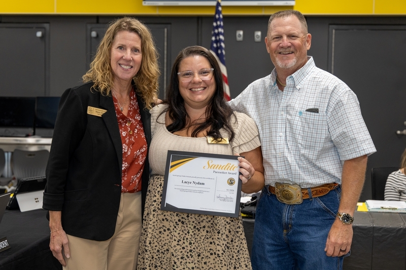 A woman holds a framed Sandite Pacesetter Award certificate while standing between two smiling colleagues.