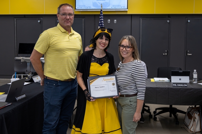 A woman in a yellow dress holds a framed Sandite Pacesetter Award certificate presented to Sherry Sanders while standing between a man in a yellow polo shirt and a woman in a striped top.