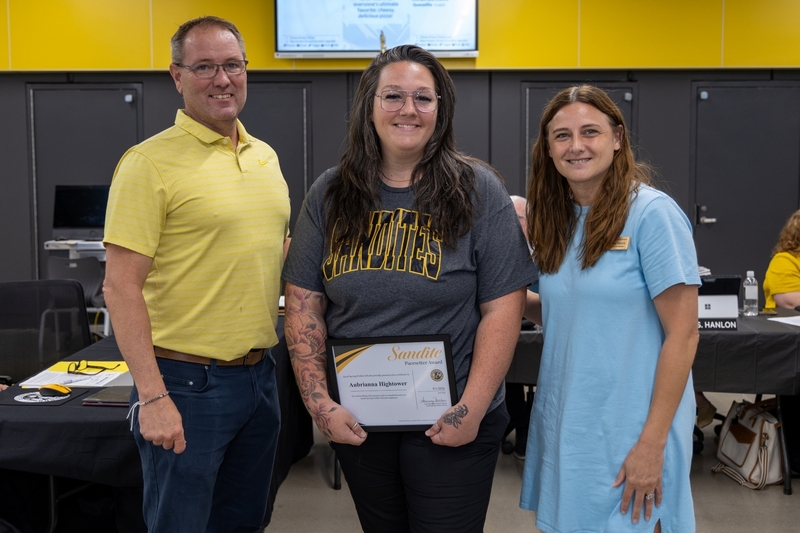 A woman holds a framed Sandite Pacesetter Award certificate while posing between two smiling colleagues.