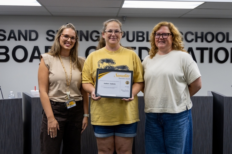 Amber Ambriz holding a framed Sandite Pacesetter Award certificate while standing between two women in front of a Sand Springs Public Schools Board of Education sign.