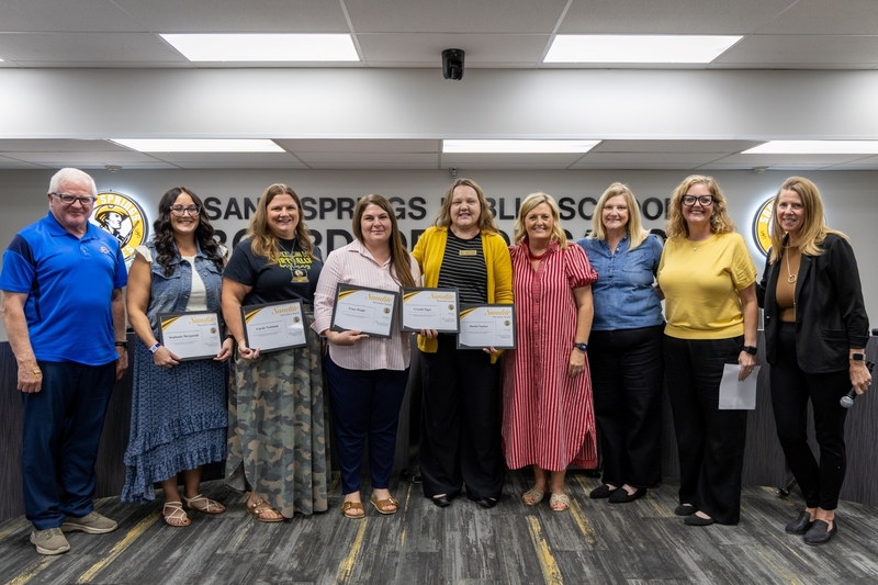 A group of four honorees and five school officials posing with framed Sandite Pacesetter Award certificates in front of the Sand Springs Public Schools Board of Education wall.