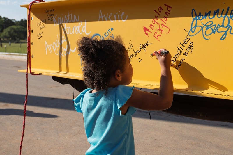 A child in a blue shirt writes on a yellow steel beam covered in signatures.