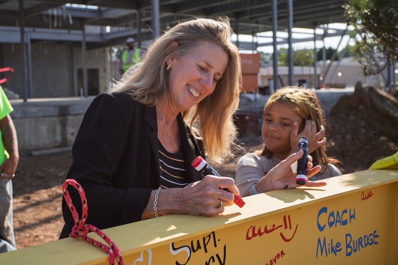 A woman smiles while signing a yellow steel beam at a construction site, with a young girl beside her.