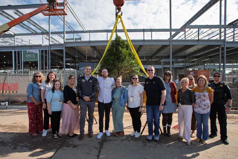 A group of people stand smiling in front of a construction site, with a small tree suspended by a steel beam and yellow straps behind them.