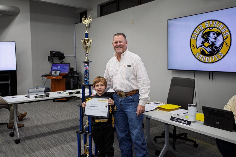 Student Nolan Vantrease holds a certificate and tall trophy while standing with board member Bo Naugle at Sand Springs Board of Education meeting, with Sandites logo displayed behind them.