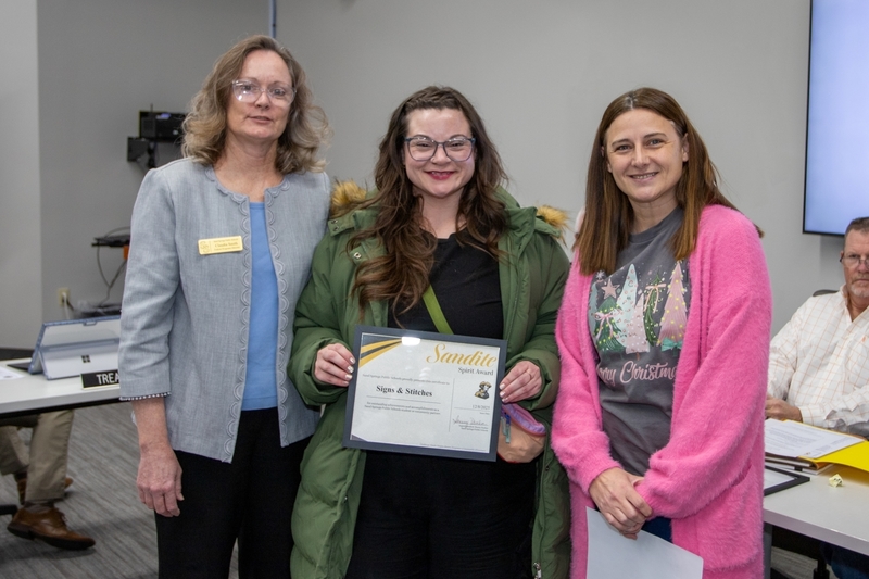 Three individuals stand together at a Board of Education meeting. The person in the center holds a Sandite Spirit Award certificate for Signs & Stitches, with two staff members standing on either side.