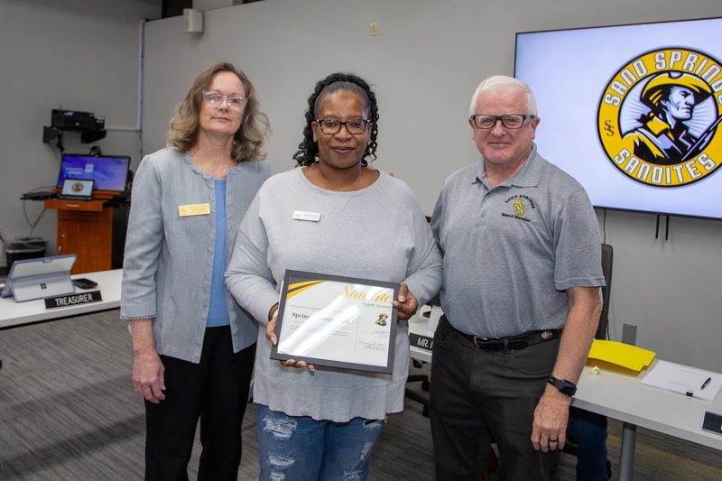Three individuals pose at a Board of Education meeting, with the center person holding a Sandite Spirit Award certificate while standing between a board member and a director.