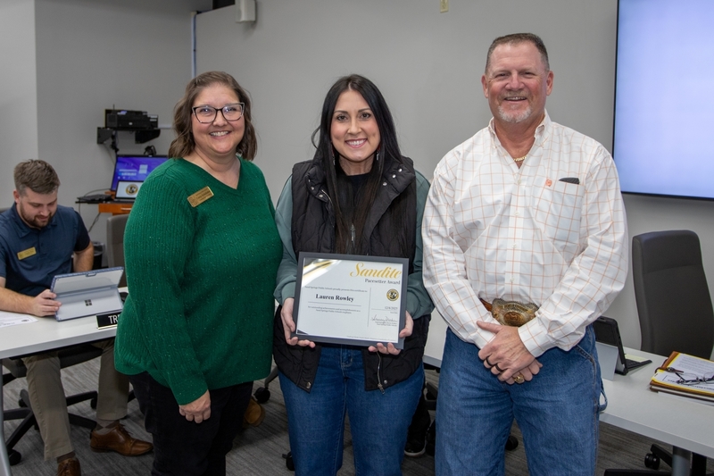 Lauren Rowley stands between two district leaders at a Board of Education meeting, holding a Sandite Pacesetter Award certificate.