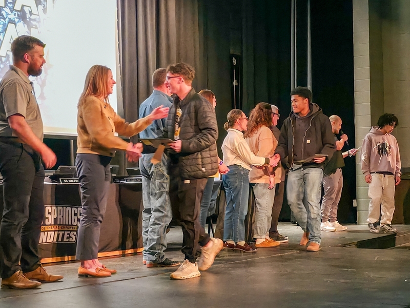 Students line up on an auditorium stage to receive certificates during a school recognition ceremony. Staff and administrators stand at the front of the stage, shaking hands and presenting awards as each student steps forward.