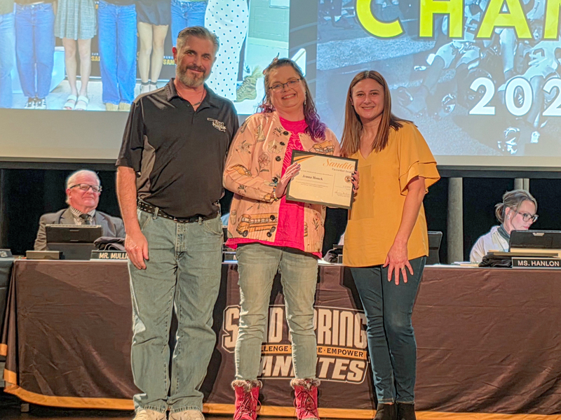 Three adults stand on a stage during a Sand Springs Public Schools awards ceremony. The person in the center holds a framed certificate and smiles while flanked by two presenters.