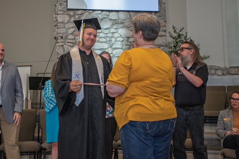 Smiling student in graduation cap and gown presents ceremonial cord to community member.
