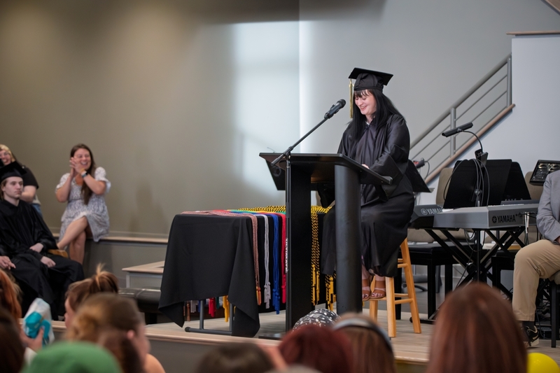 Student in cap and gown stands at lectern on stage and addresses the crowd.