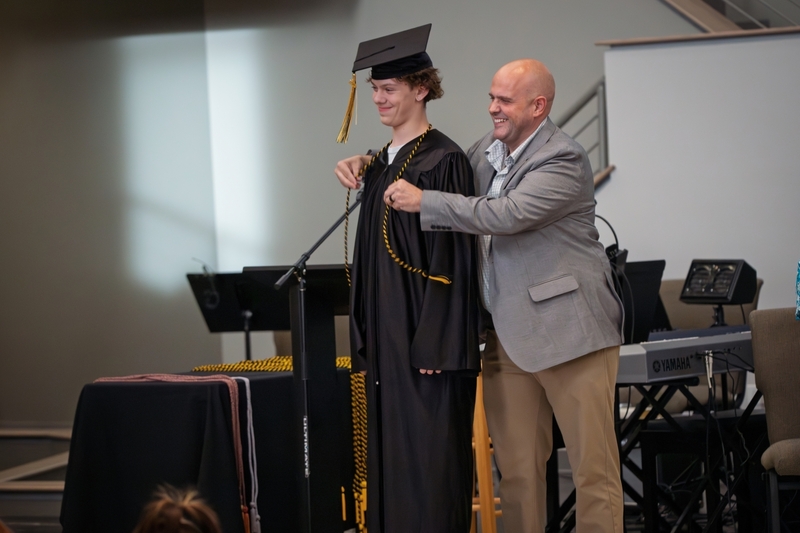 Principal Jay Rotert places ceremonial cord on student in graduation cap and gown.