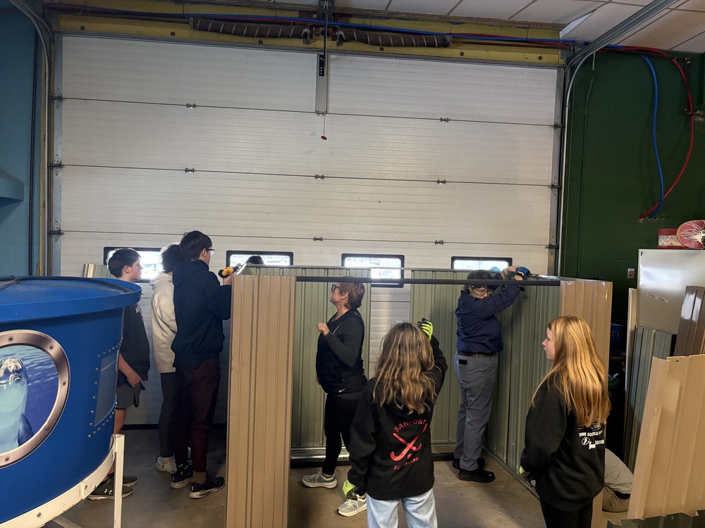 When Sanford Middle School 7th graders returned from the Ecology School this fall with ideas about making outdoor time more engaging, the teachers listened and students are now building the solution themselves. Students in the Environmental Club and Mechanical Advantage Clubs are assembling a storage shed funded by the Stronger Connections Committee grant, creating a resource that will benefit the entire school community while gaining valuable hands-on construction experience. The project began with student feedback. During their week-long trip to the Ecology School, 7th graders experienced outdoor activities like volleyball, giant Jenga, and oversized Connect Four that kept them active and engaged. Diana Allen, who serves on the Stronger Connections Committee with colleague Ms. Mason, recognized an opportunity to translate student input into action. "We have this space out here that's not going to be effective for use during flex time," Allen noted during a recent presentation to students. "Instead of just walking around in circles around the building... how can we make that better for all of us?" Rather than simply purchasing and installing equipment, the school turned the project into a learning opportunity. Students from two clubs joined forces to construct the shed that would house the new outdoor equipment. Ms. Mason's Mechanical Advantage Club has been building giant Jenga blocks from two-by-fours, while both clubs have worked together on shed assembly. Allen acknowledged the collaborative effort. "Ms. Mason and I are on the Stronger Connections committee. We got permission to put it outside. And we are using the Environmental Club and Mechanical Advantage Club to put the shed together," she said. The hands-on work has resonated with students across grade levels. "Building the shed is really fun. I like the hands on. It's also a good idea and I like helping the community," said Owen Jutras, an 8th grader in the Environmental Club. Jediah Bowden, also in 8th grade, reflected on the project's lasting impact: "I am very happy to be a part of building the shed to store equipment so we can have fun outside during FLEX time. And the school can enjoy it for years to come!" Fellow 8th grader Eamon McGuckin Welsch added, "I have found the shed building to be extremely rewarding, finding it to be a great use of our time and resources to make something for the school." Sixth grader Jordan Barron appreciated the practical skills: "It's been fun, I really liked putting on the siding with a screwdriver." The shed will soon be stocked with volleyball nets, giant games, and PTA-donated activity totes, ready to transform how students experience outdoor time at Sanford Middle School.