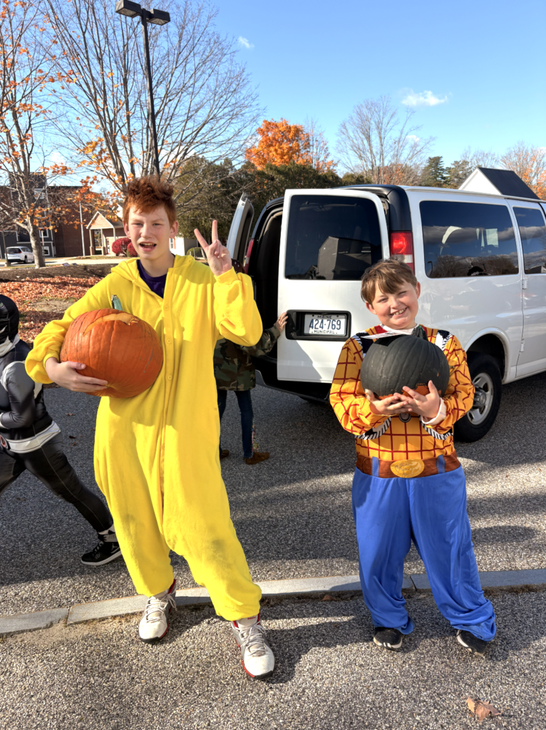 Two students pose with pumpkins outside of a van.