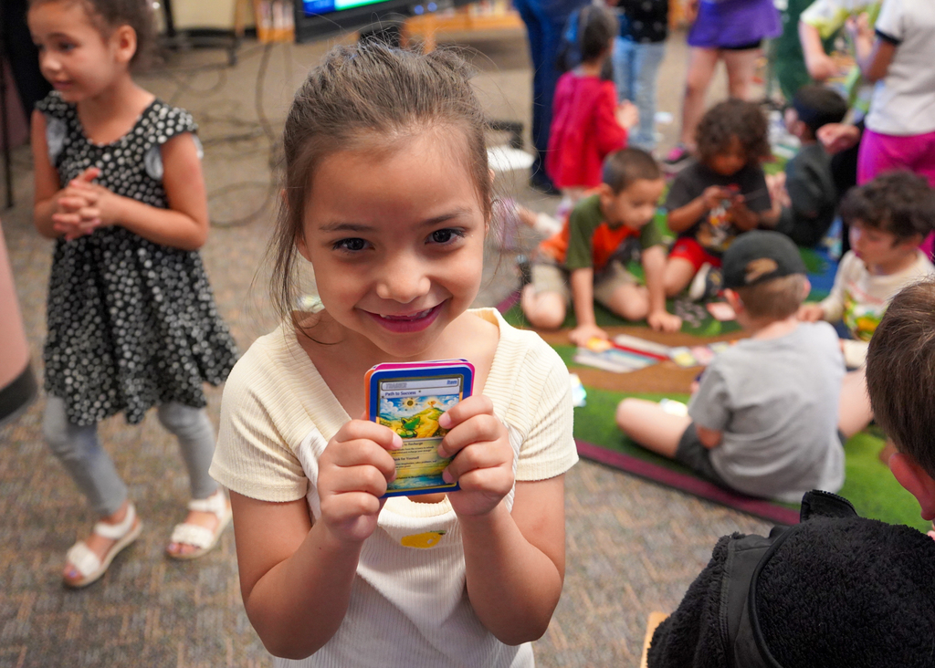 A young girl holds a card in both hands and smiles. Behind her, children sit on a rug. Some are engaged in activities.
