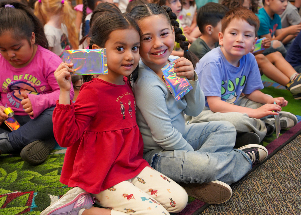 A group of young children, seated on a carpeted floor, holds up colorful objects. One child wears red and white pajamas.