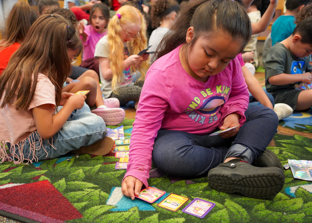 Children sit on a rug, focusing on playing a card game. A girl in pink looks down while holding cards.