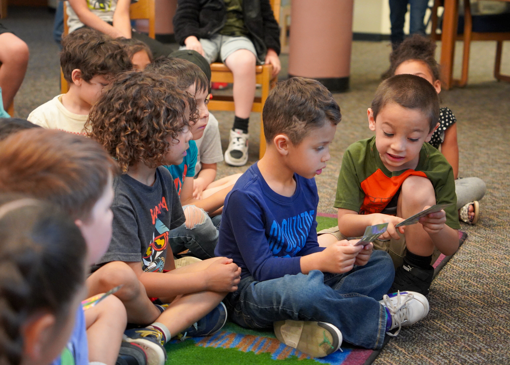 A group of children sits on the floor, focusing on a tablet held by one child. They are in a carpeted area.