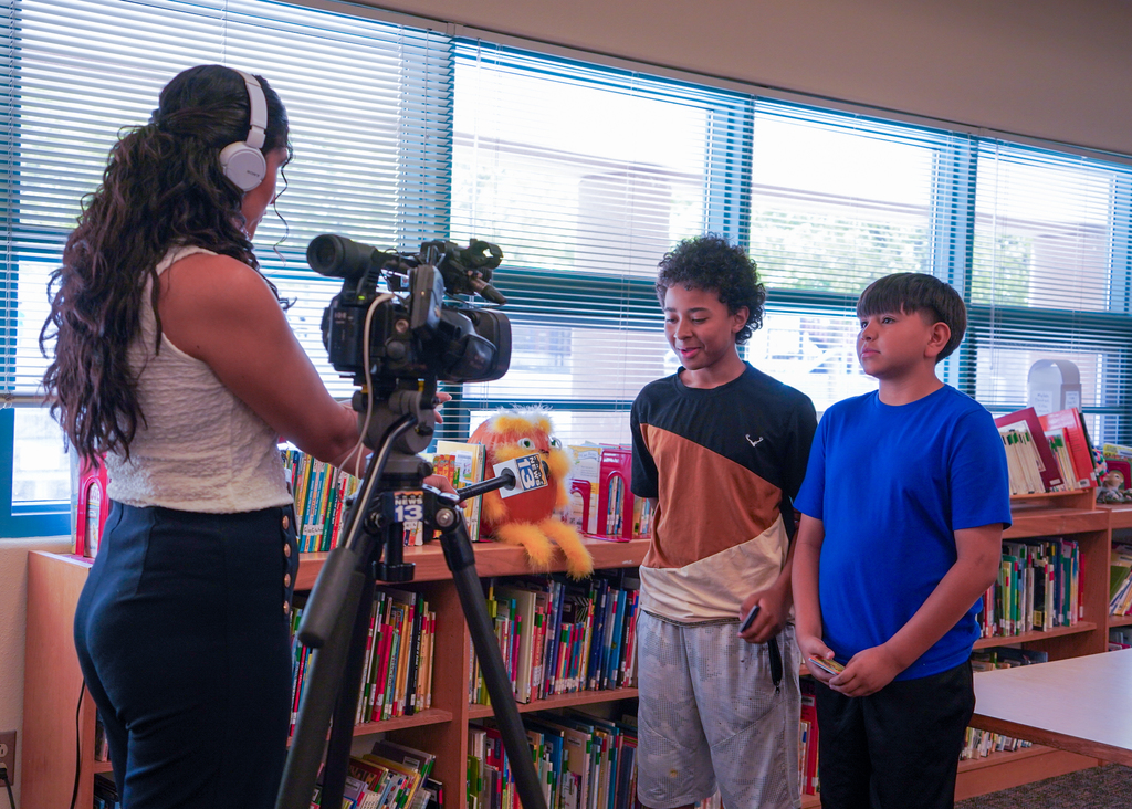 A woman with headphones films two boys with a camera on a tripod in a library.