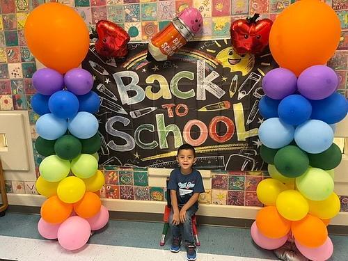A child sits in front of a colorful balloon arch and a chalkboard that says "Back to School".