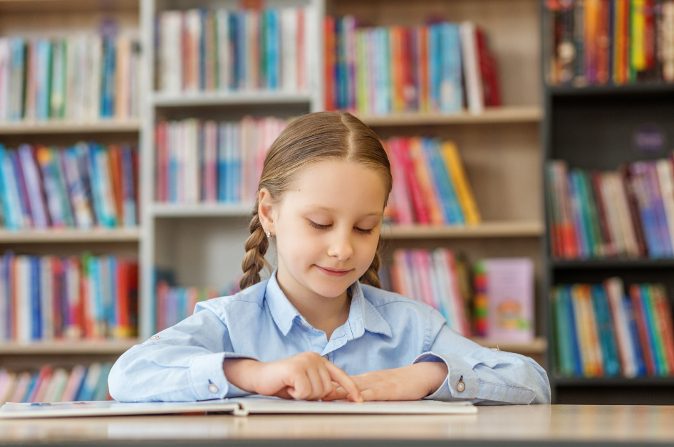 A girl with braided hair sits at a desk in a library. She reads a book with both hands.