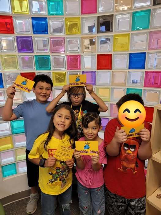 A group of children  hold yellow cards with smiley faces in front of a multicolored wall.