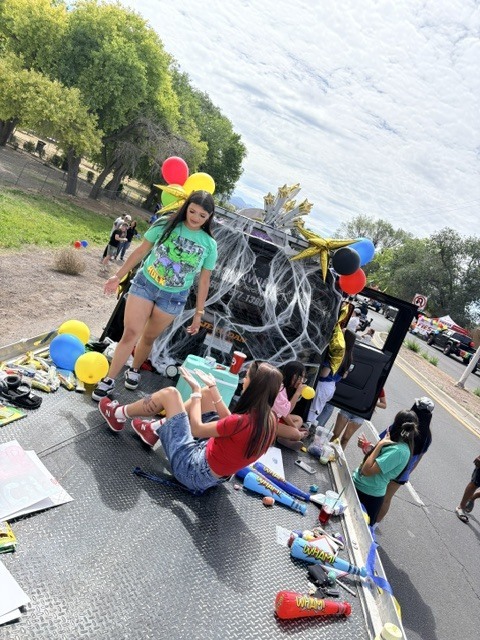 People on a trailer with balloons, decorations, and toys in a street with trees.