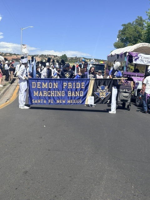 A marching band holds a banner reading "Demon Pride Marching Band, Santa Fe, New Mexico" in a parade.