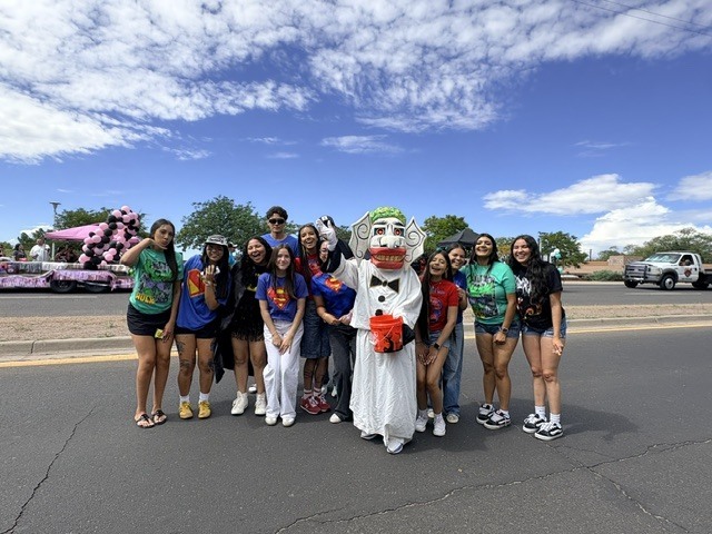Group of people in a parade, standing on road, with a clown mascot in white costume.