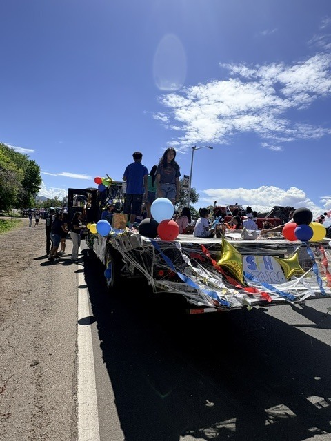 Parade float with people on it, adorned with colorful balloons and banners, moving on a road.