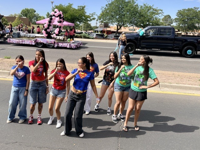 Seven women in various clothing stand in a street, smiling, with a black truck and a float in the background.